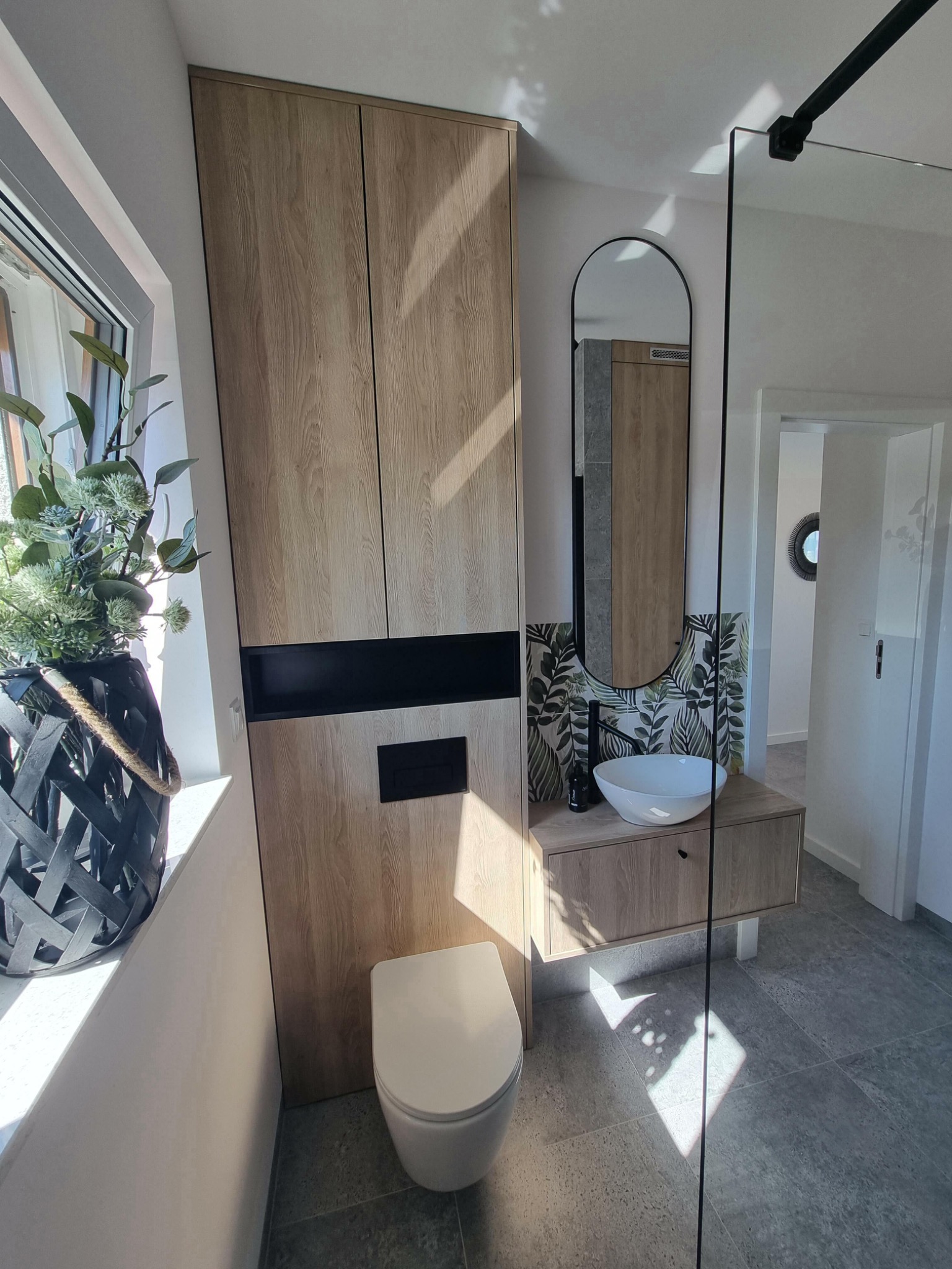 Nowoczesna łazienka z zabudową meblową z drewnopodobnymi frontami, wisząca umywalka z szafką, lustro w czarnej ramie i szklana kabina prysznicowa.