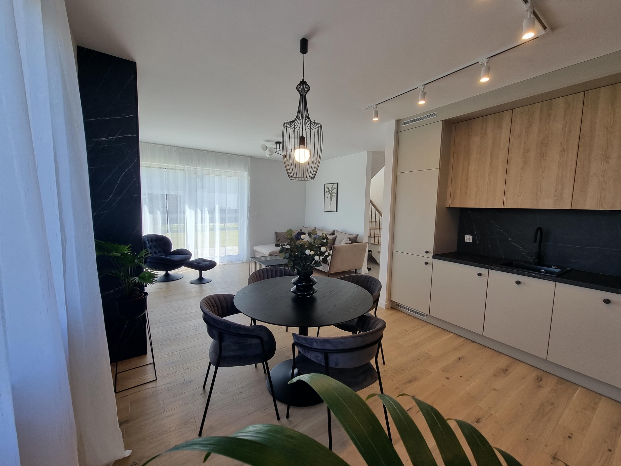 Jasne wnętrze z okrągłym, czarnym stołem i welurowymi krzesłami, widok na salon z szarą sofą i fotelami, drewniana podłoga, zabudowa kuchenna z drewnianymi frontami i czarnym blatem, czarna lampa...
