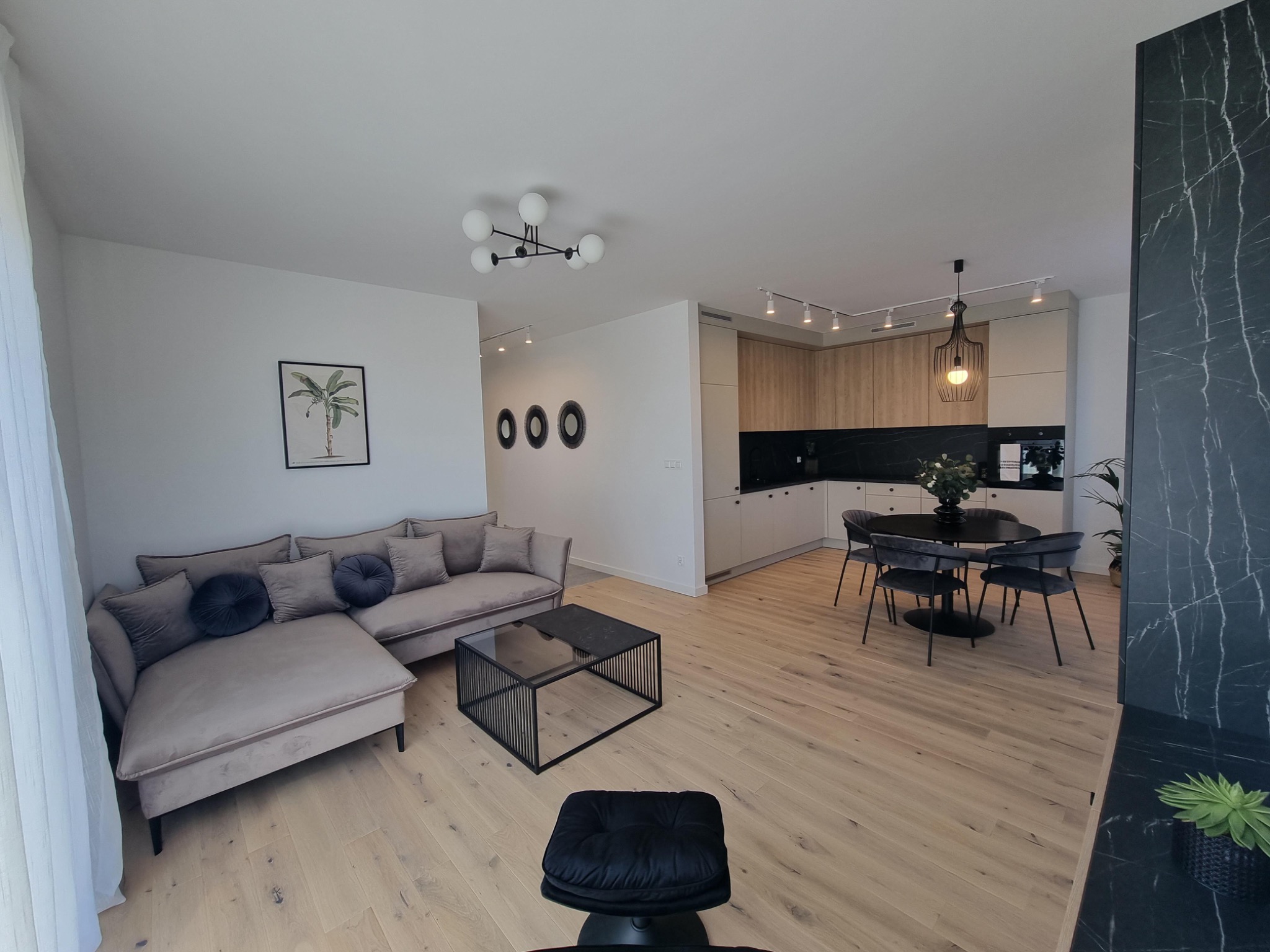 Przestronne wnętrze z otwartą kuchnią, beżowa sofa z okrągłymi poduszkami, stolik kawowy z czarną ramą, okrągły stół z krzesłami w części jadalnej, drewniana podłoga i zabudowa kuchenna...