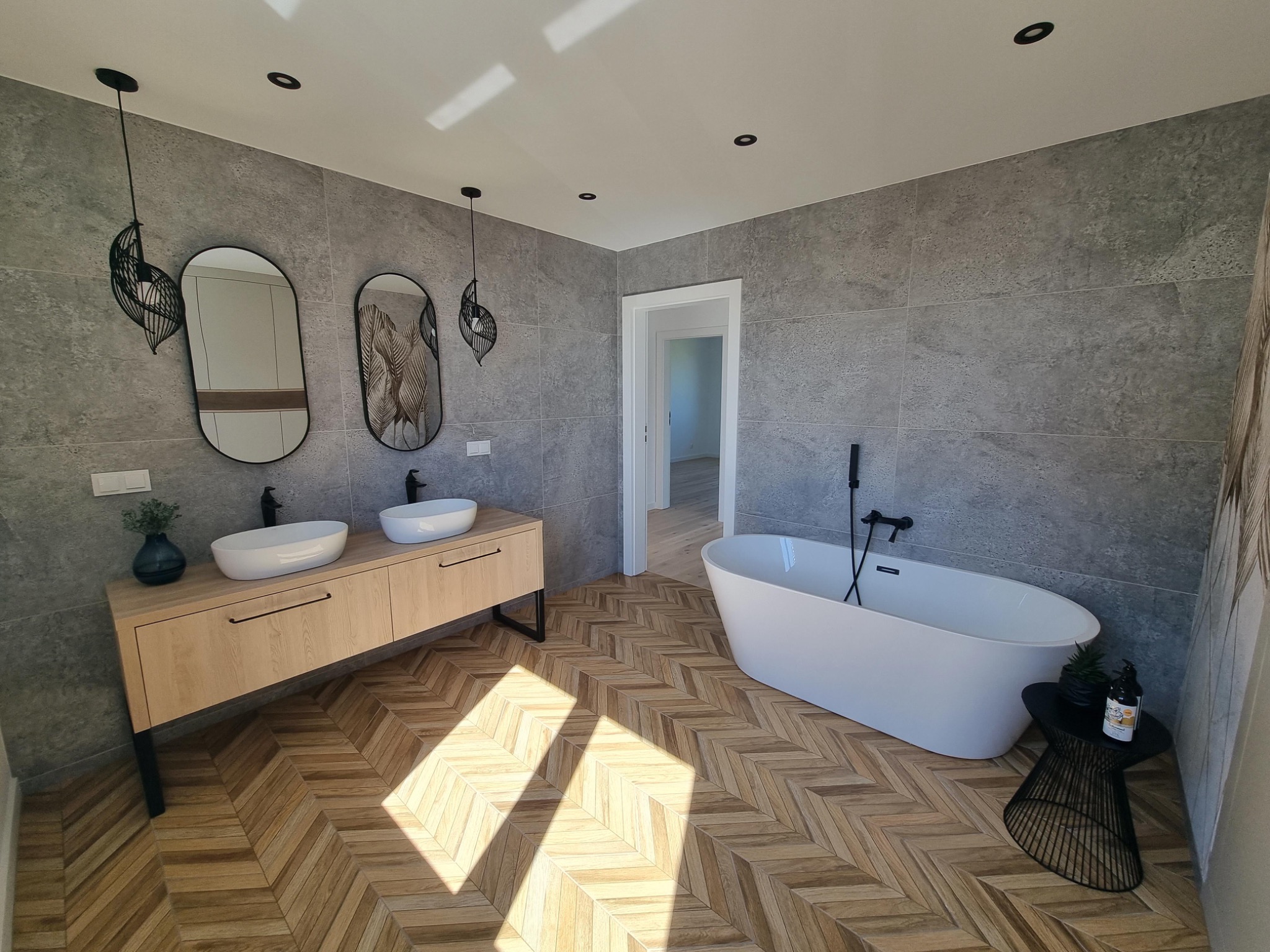 Nowoczesna łazienka z drewnianą podłogą ułożoną w jodełkę, szare płytki ścienne, podwójna umywalka na drewnianej szafce, wolnostojąca biała wanna z czarną armaturą i dekoracyjne lampy wiszące.
