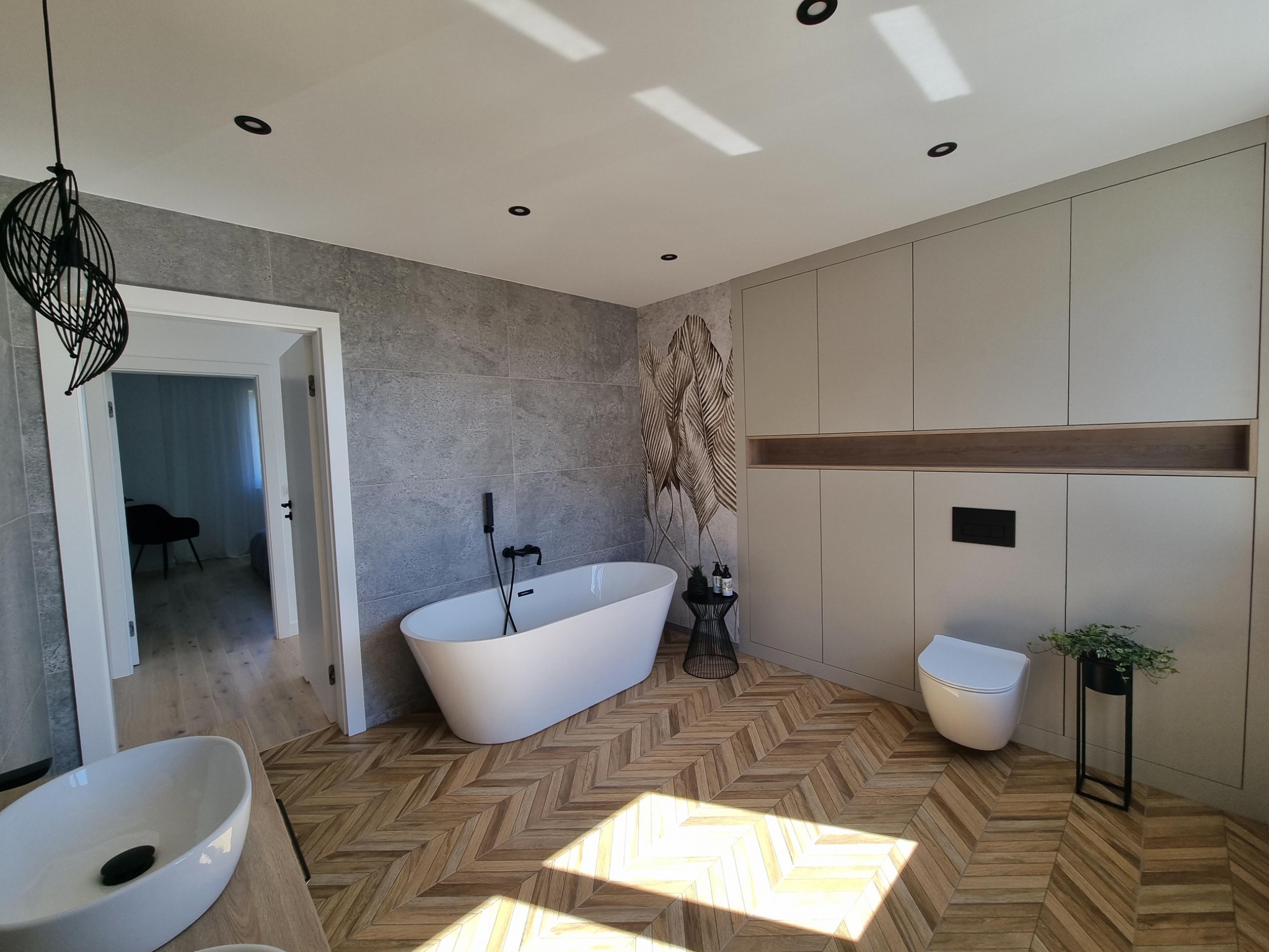 Nowoczesna łazienka z wolnostojącą wanną, umywalką nablatową, toaletą i zabudową meblową w odcieniach szarości, z drewnianą podłogą ułożoną w jodełkę i designerską lampą wiszącą.