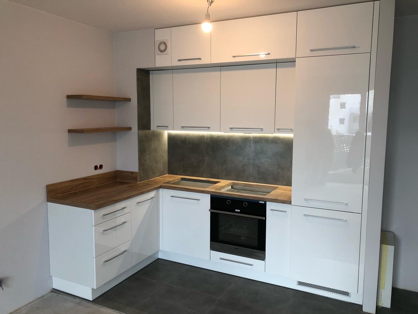 Nowoczesna, biała kuchnia z drewnianymi blatami, szarym backsplashiem, oświetleniem LED podszafkowym i dwoma drewnianymi półkami na ścianie. Widoczny piekarnik i płyta grzewcza. Stan surowy...