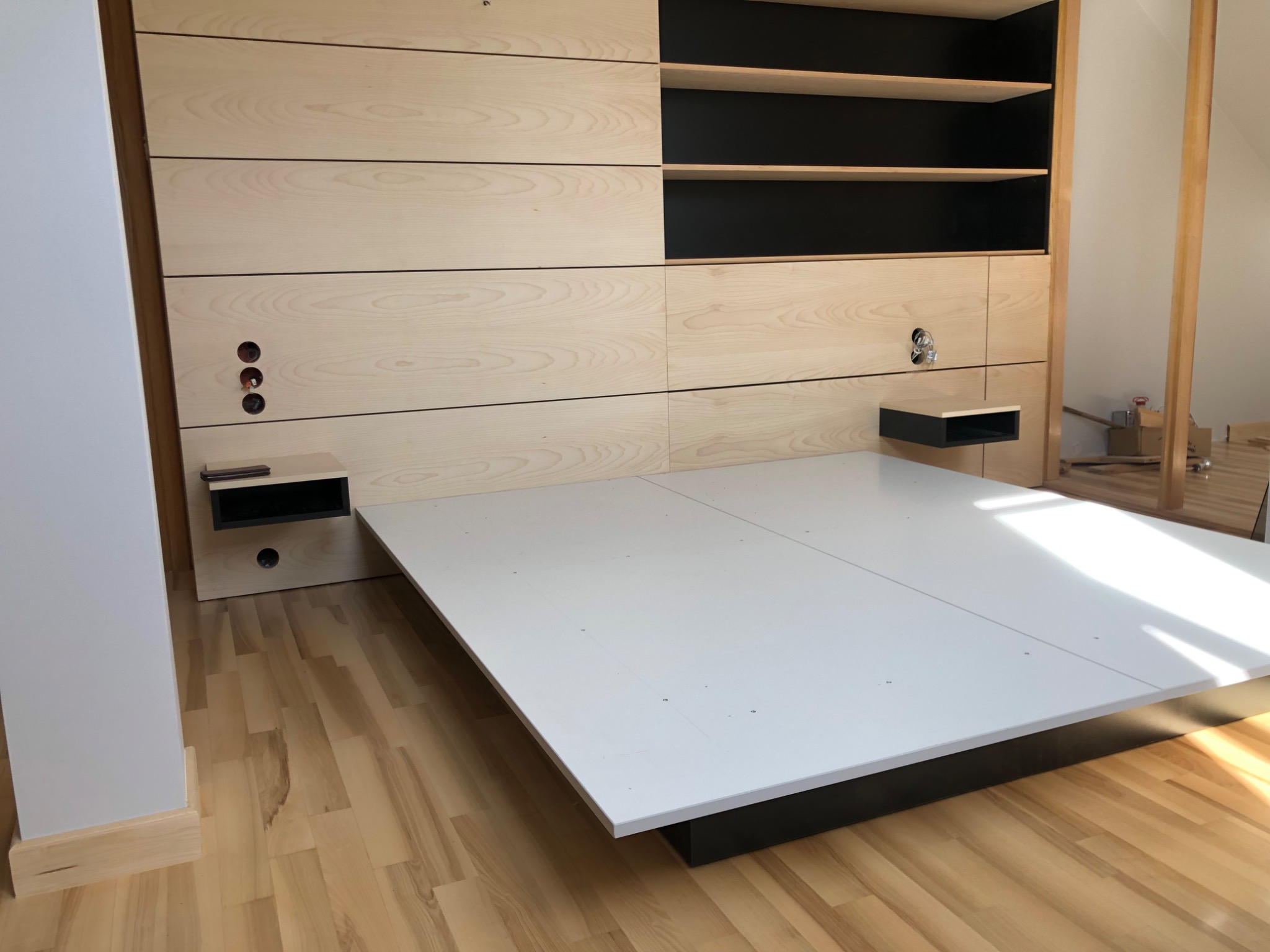 Montaż łóżka na wymiar z drewnianym panelem ściennym i szafkami nocnymi w kolorze czarnym, widoczne otwory na gniazdka elektryczne.