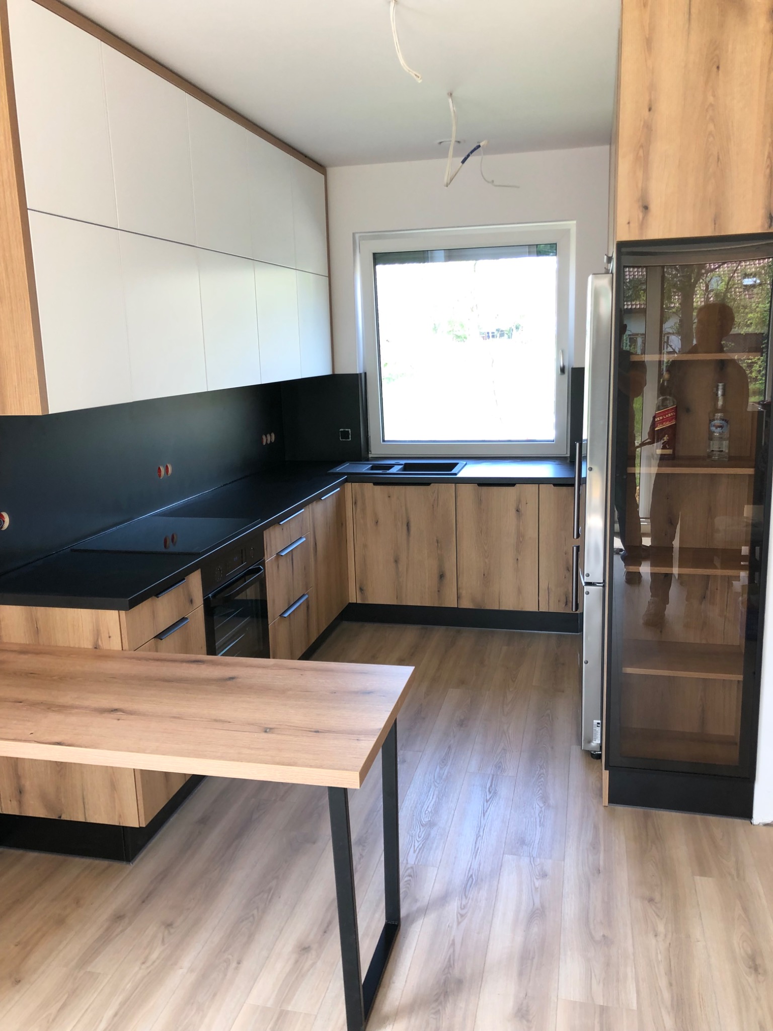 Nowoczesna kuchnia z drewnianymi frontami szafek, czarnym blatem i panelem ściennym, białymi szafkami górnymi, oraz wyspą z drewnianym blatem na metalowych nogach. Widoczna przeszklona lodówka...