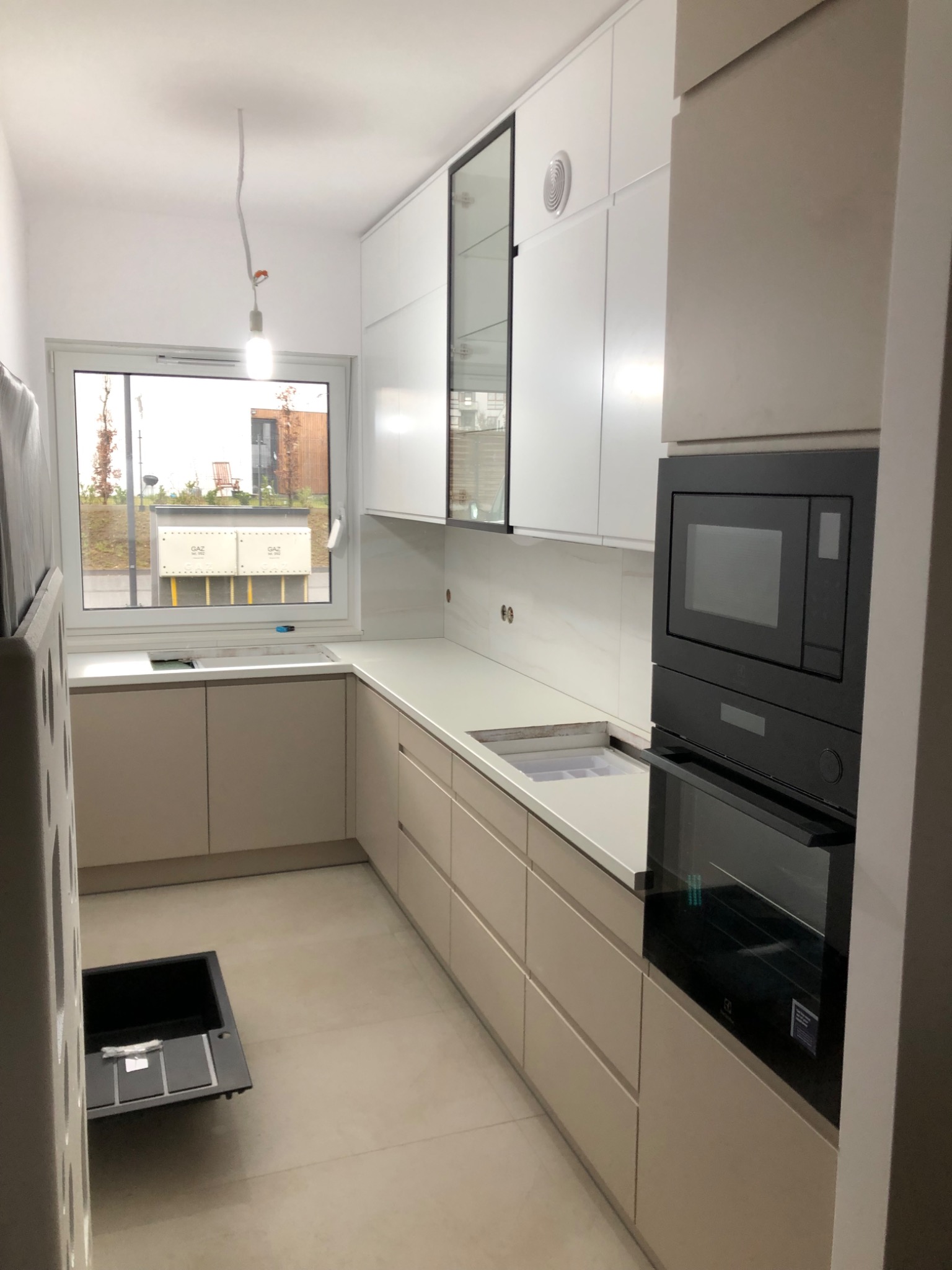 Wnętrze kuchni w trakcie montażu z beżowymi frontami szafek, białym blatem, zabudowaną czarną mikrofalówką i piekarnikiem, oraz widocznym oknem z widokiem na zewnątrz i niezabezpieczoną żarówką.