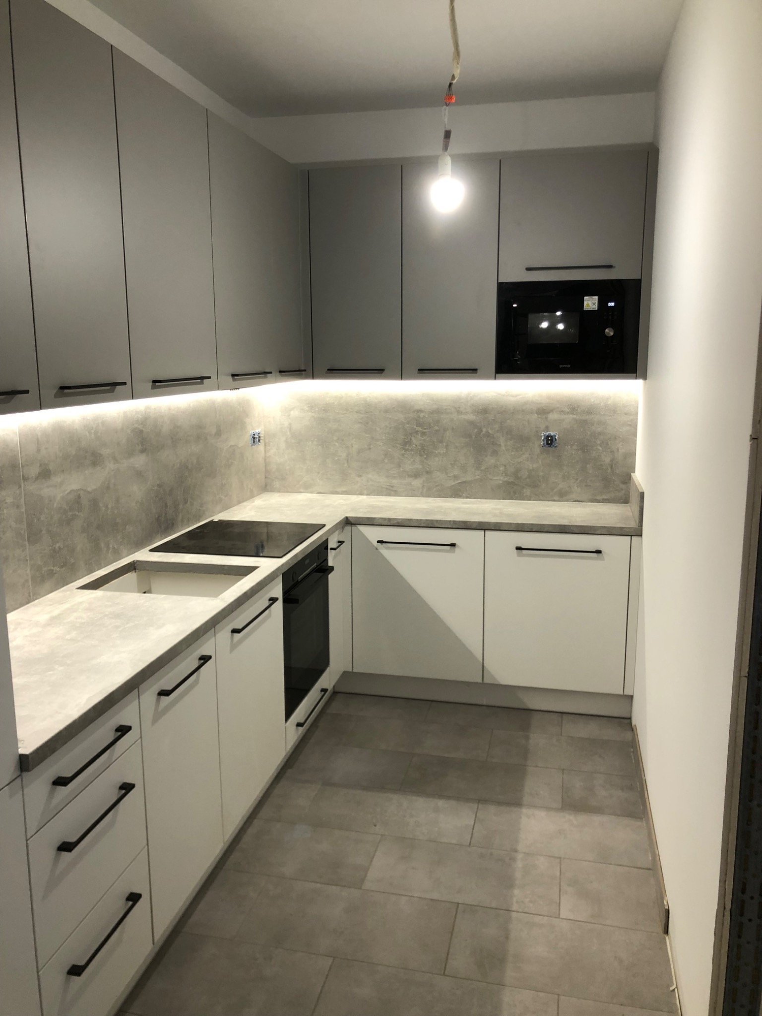 Nowoczesna kuchnia z szarymi górnymi szafkami i białymi dolnymi, czarne długie uchwyty, szary blat, podświetlenie LED pod szafkami, płyta indukcyjna i piekarnik w zabudowie.