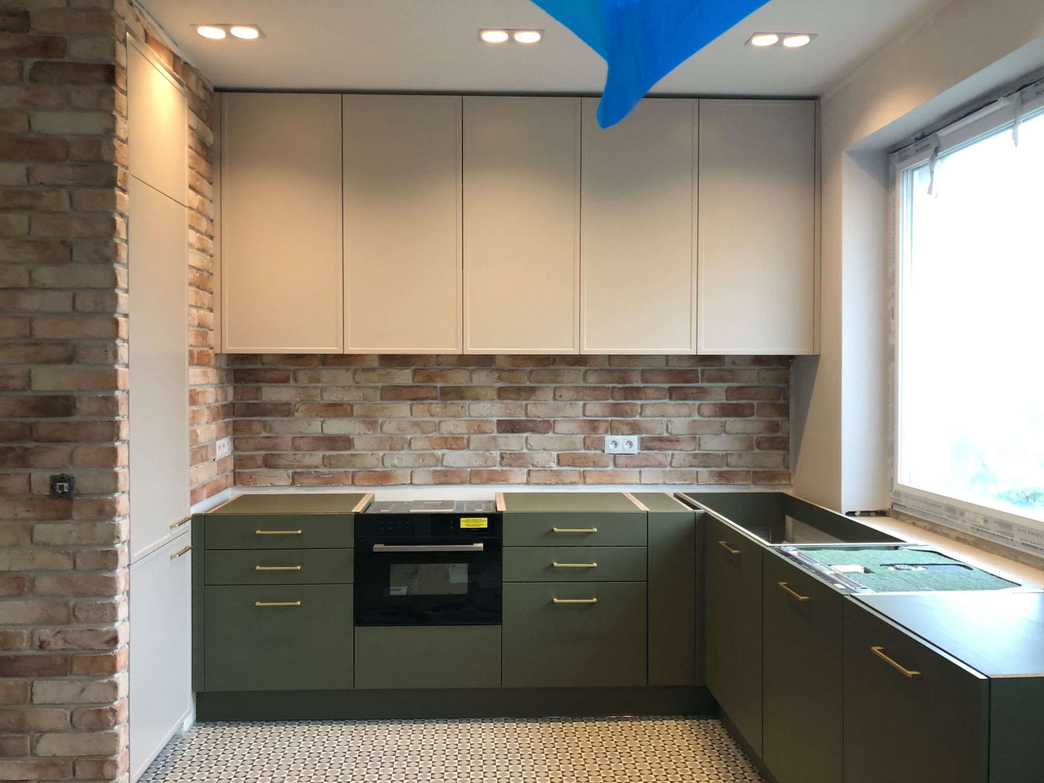 Nowoczesna kuchnia z oliwkowymi szafkami dolnymi i białymi górnymi, ceglana ściana nad blatem i wzorzysta podłoga w trakcie montażu.