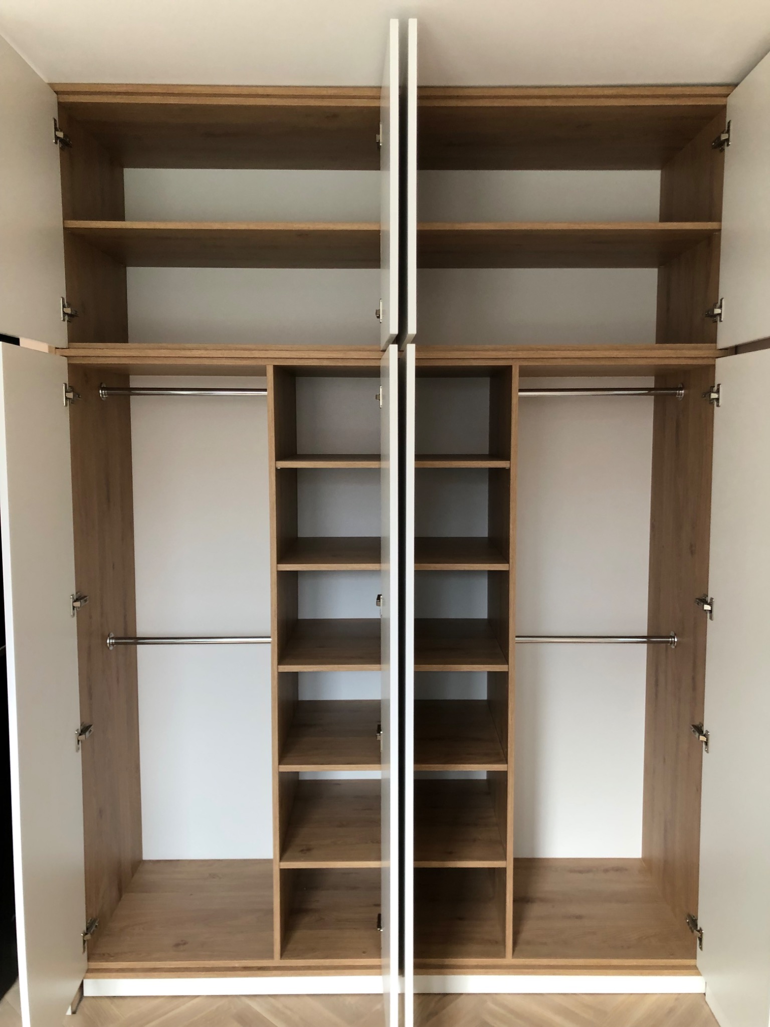 Wnętrze szafy wnękowej z otwartymi drzwiami, widoczne półki w kolorze drewna i metalowe drążki na ubrania, białe ściany wewnątrz.