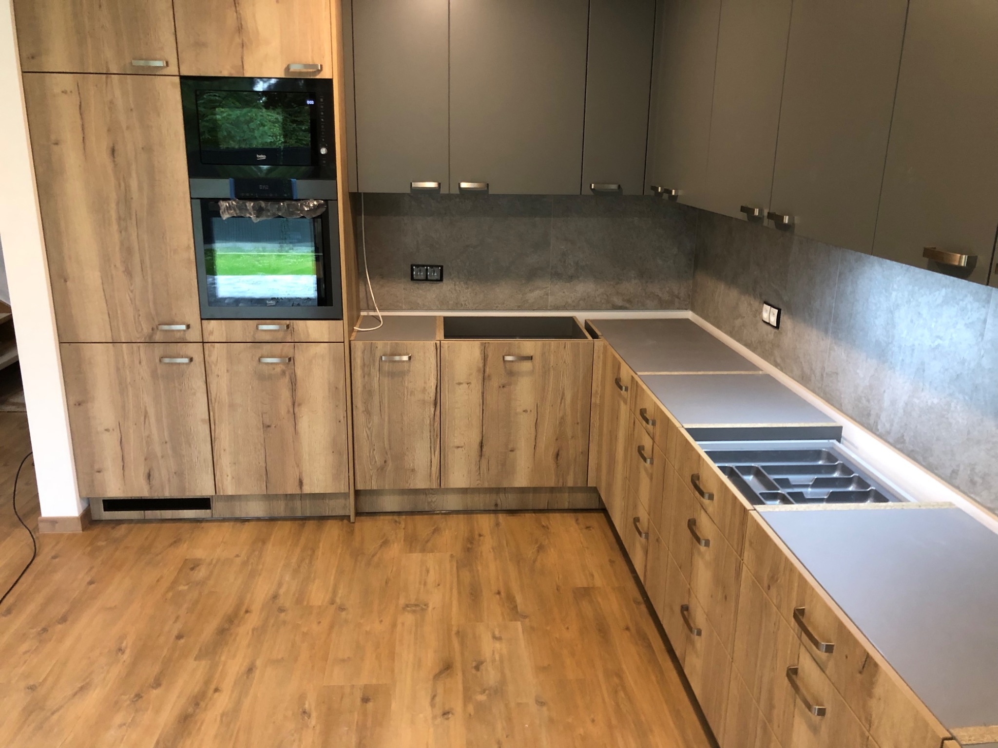 Nowoczesna kuchnia na wymiar z drewnianymi frontami i szarymi górnymi szafkami, widoczny piekarnik i płyta grzewcza, otwarte szuflady z wkładem na sztućce.