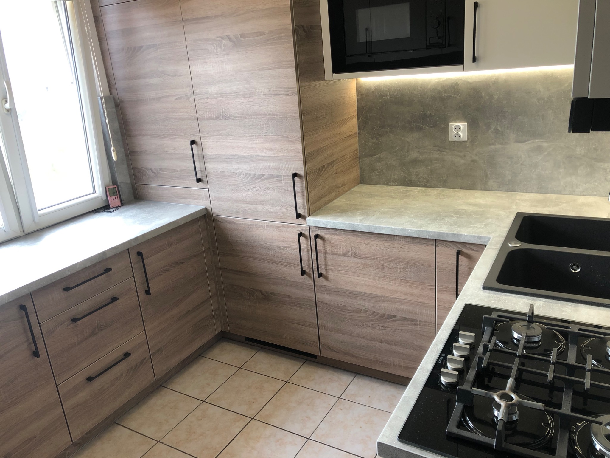 Jasna kuchnia z drewnopodobnymi frontami szafek i szarym blatem, widoczna płyta gazowa i okno z roletą.