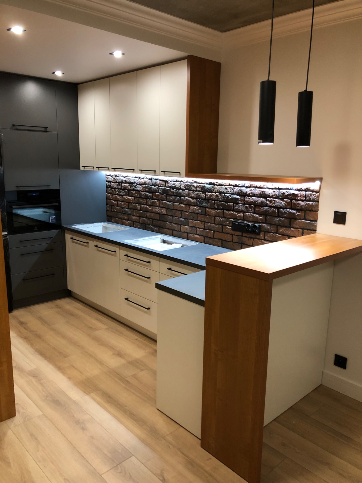 Nowoczesna kuchnia z białymi i szarymi szafkami, blatem z szarego kamienia i ścianą z cegły podświetloną od dołu, uzupełniona drewnianym blatem barowym i minimalistycznymi czarnymi lampami wiszącymi.