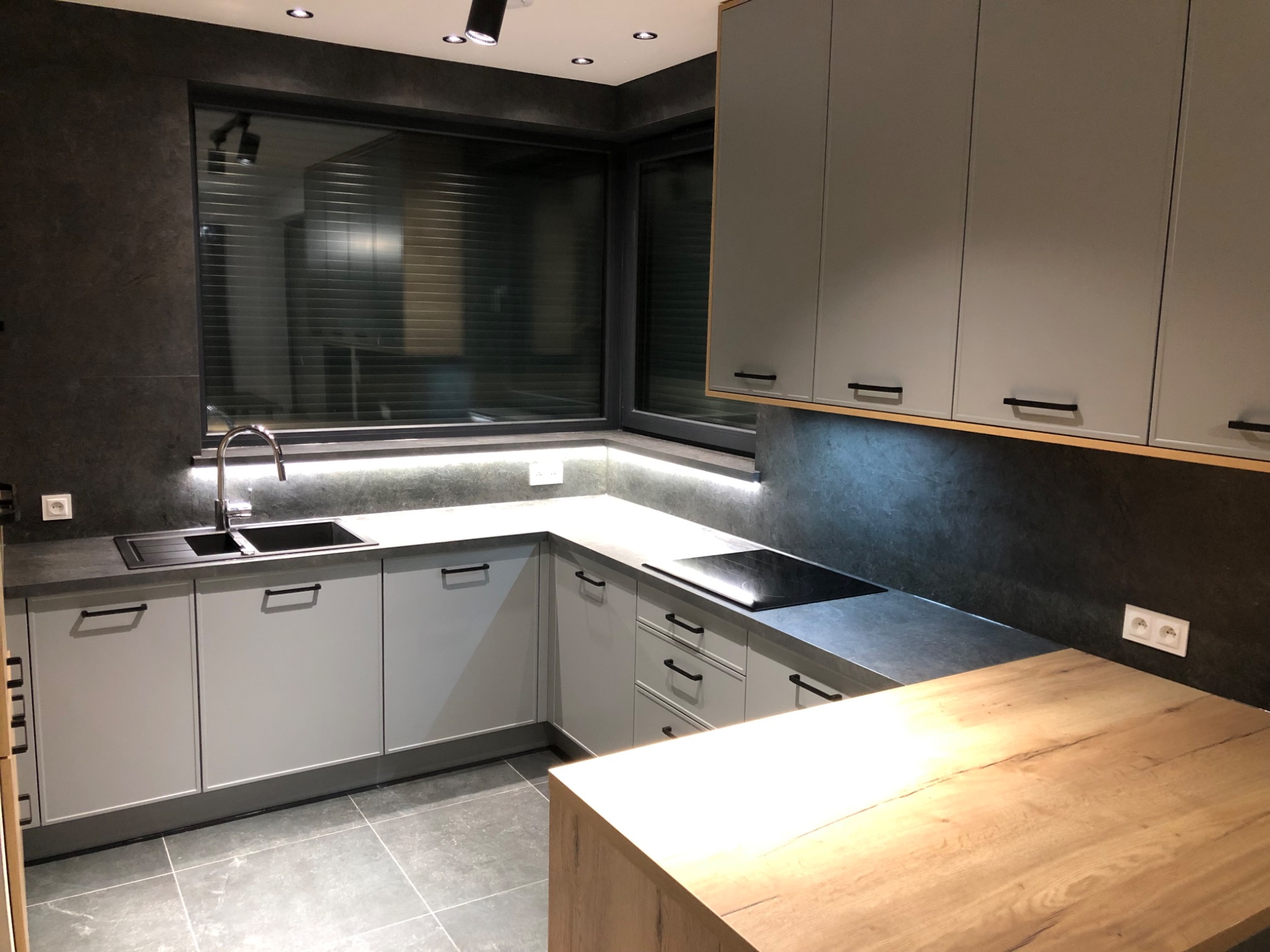 Nowoczesna kuchnia w szarościach z drewnianym blatem wyspy, czarnym zlewem i płytą indukcyjną, oświetlona ledowym pasem pod szafkami wiszącymi.