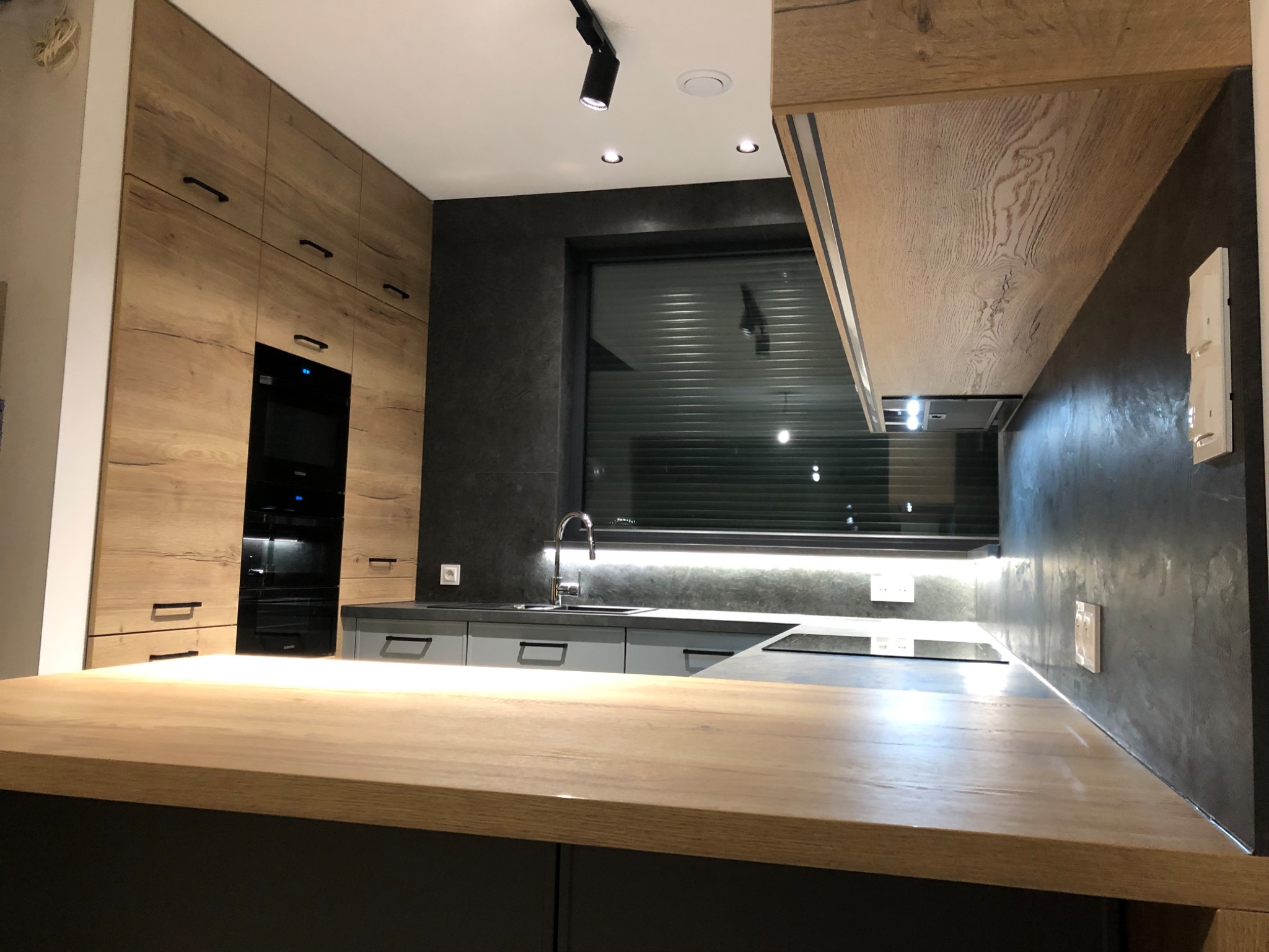 Nowoczesna kuchnia z drewnianymi frontami i blatem, szare szafki dolne, czarne ściany i okno z żaluzjami, oświetlenie punktowe.