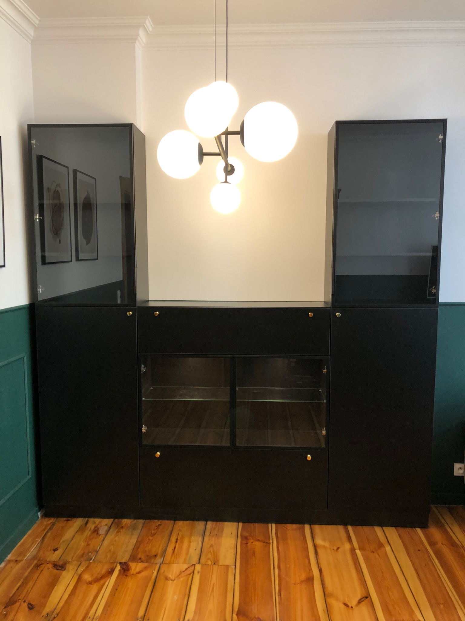 Czarna zabudowa meblowa z przeszklonymi witrynami i złotymi gałkami, oświetlona nowoczesną lampą kulistą we Wrocławiu, na tle parkietu i ściany w dwóch odcieniach.