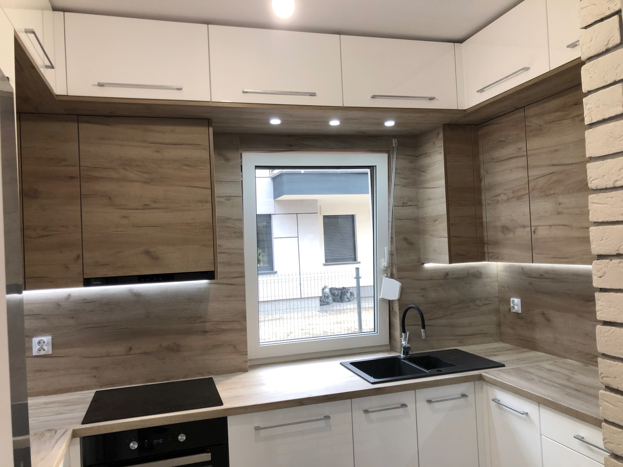 Nowoczesna kuchnia z białymi szafkami dolnymi i górnymi z drewnianymi frontami, czarny zlew pod oknem, ceglana ściana po prawej stronie.