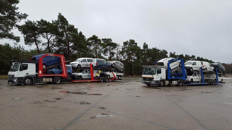 Dwa autotransportery Mercedes, załadowane samochodami osobowymi i vanami, stoją na parkingu w pochmurny dzień. Widoczne drzewa w tle.