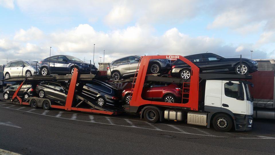 Czerwono-biała laweta Lohr przewozi różnokolorowe samochody osobowe, widok z boku, pochmurne niebo w tle. Transport aut z zagranicy.