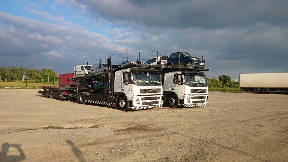 Dwa autotransportery Volvo, jeden czarny, drugi biały, załadowane samochodami osobowymi, stoją na parkingu pod pochmurnym niebem.