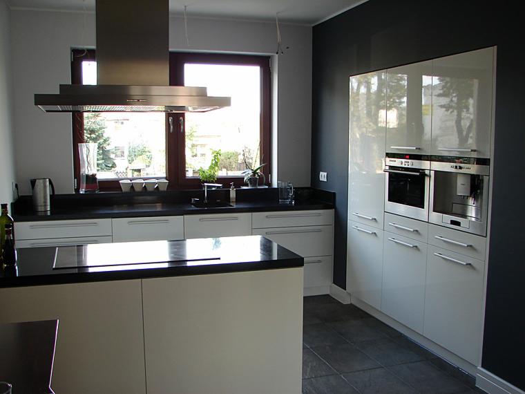 Nowoczesna kuchnia z białymi, lakierowanymi frontami, czarnym blatem i wyspą kuchenną, zintegrowanym piekarnikiem i ekspresem do kawy.