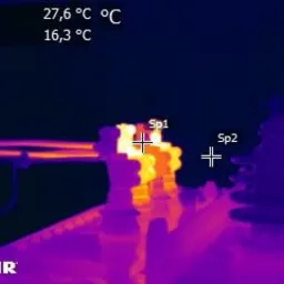 Obraz termowizyjny transformatora średniego napięcia z zaznaczonymi punktami pomiarowymi Sp1 i Sp2. Punkt Sp1, po przeczyszczeniu styków, wskazuje temperaturę 27,6°C, natomiast Sp2 – 16,3°C.