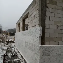 Budowa domu w zimowej scenerii: ściany z białych bloczków z widocznymi otworami okiennymi i częściowo nałożonymi płytami izolacyjnymi z widoczną strukturą, szara, pochmurna pogoda.