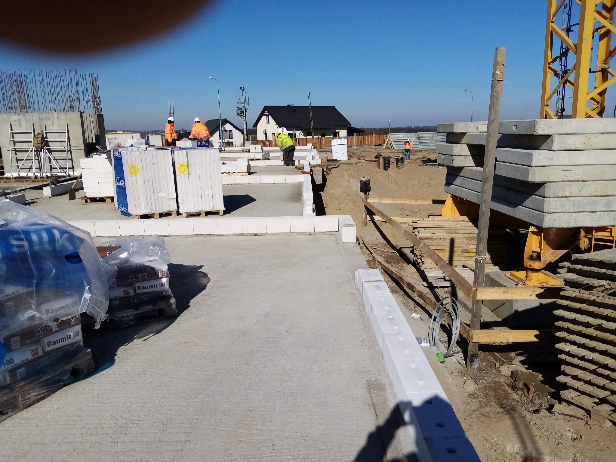 Budowa domu jednorodzinnego: fundamenty, bloczki Silka, materiały budowlane Baumit, pracownicy w odblaskowych kamizelkach, żuraw budowlany w tle, słoneczny dzień.