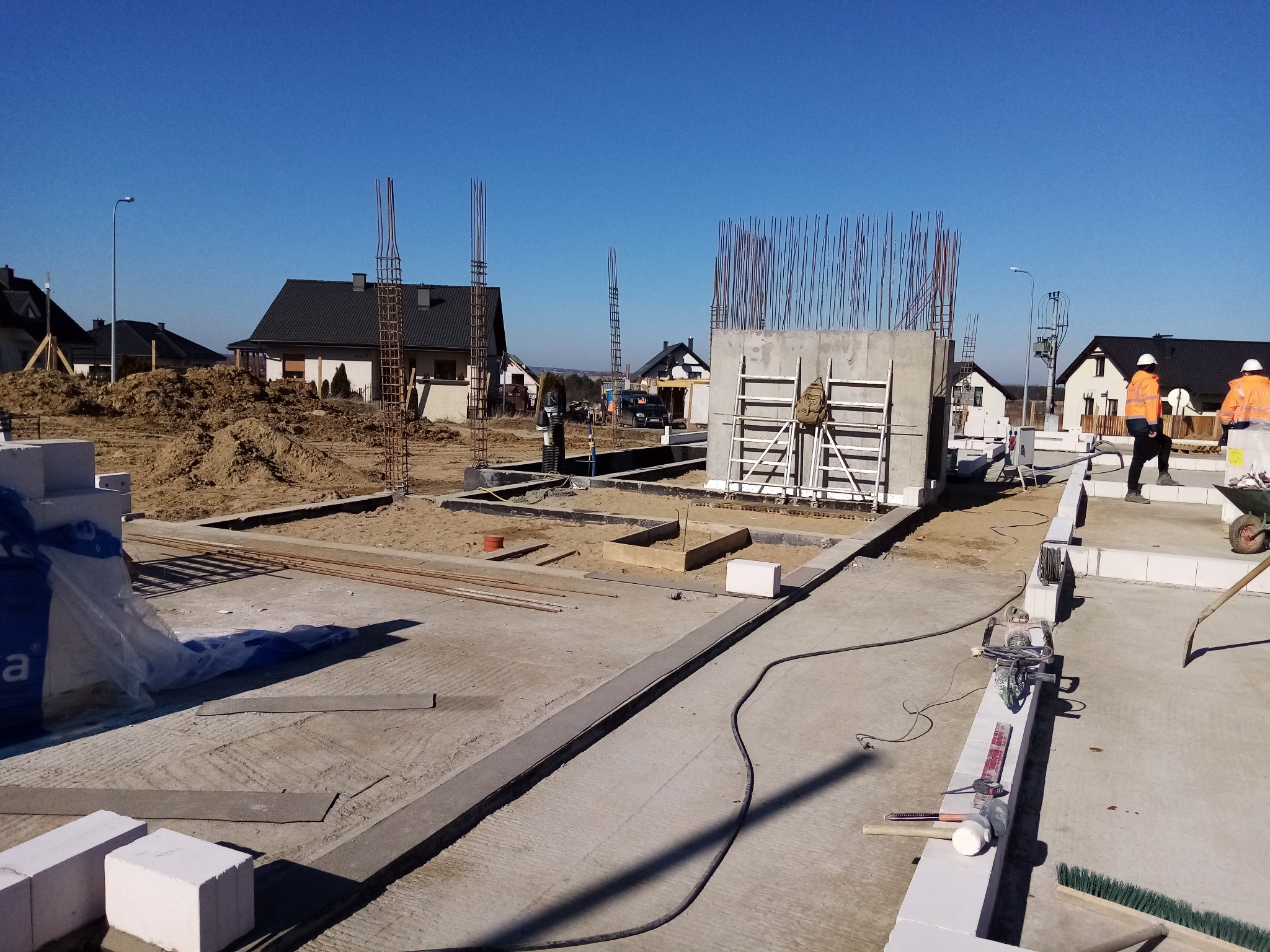 Budowa domu jednorodzinnego: fundamenty, zbrojenie, betonowe ściany szczytowe, pracownicy w pomarańczowych kurtkach, błękitne niebo w tle.