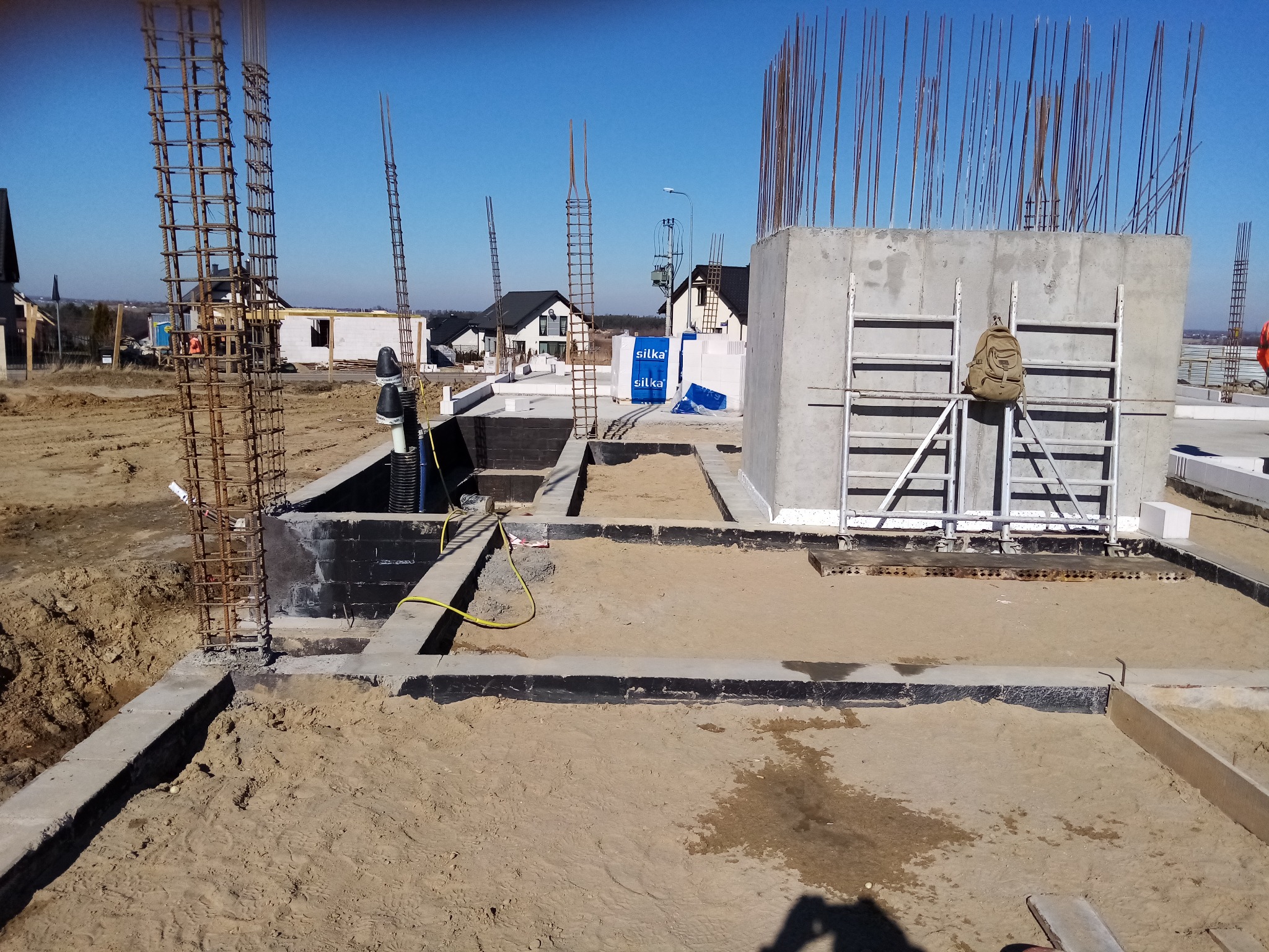 Fundamenty domu jednorodzinnego w trakcie budowy, widoczne zbrojenia, izolacja przeciwwilgociowa, piasek zasypowy oraz drabina z plecakiem.
