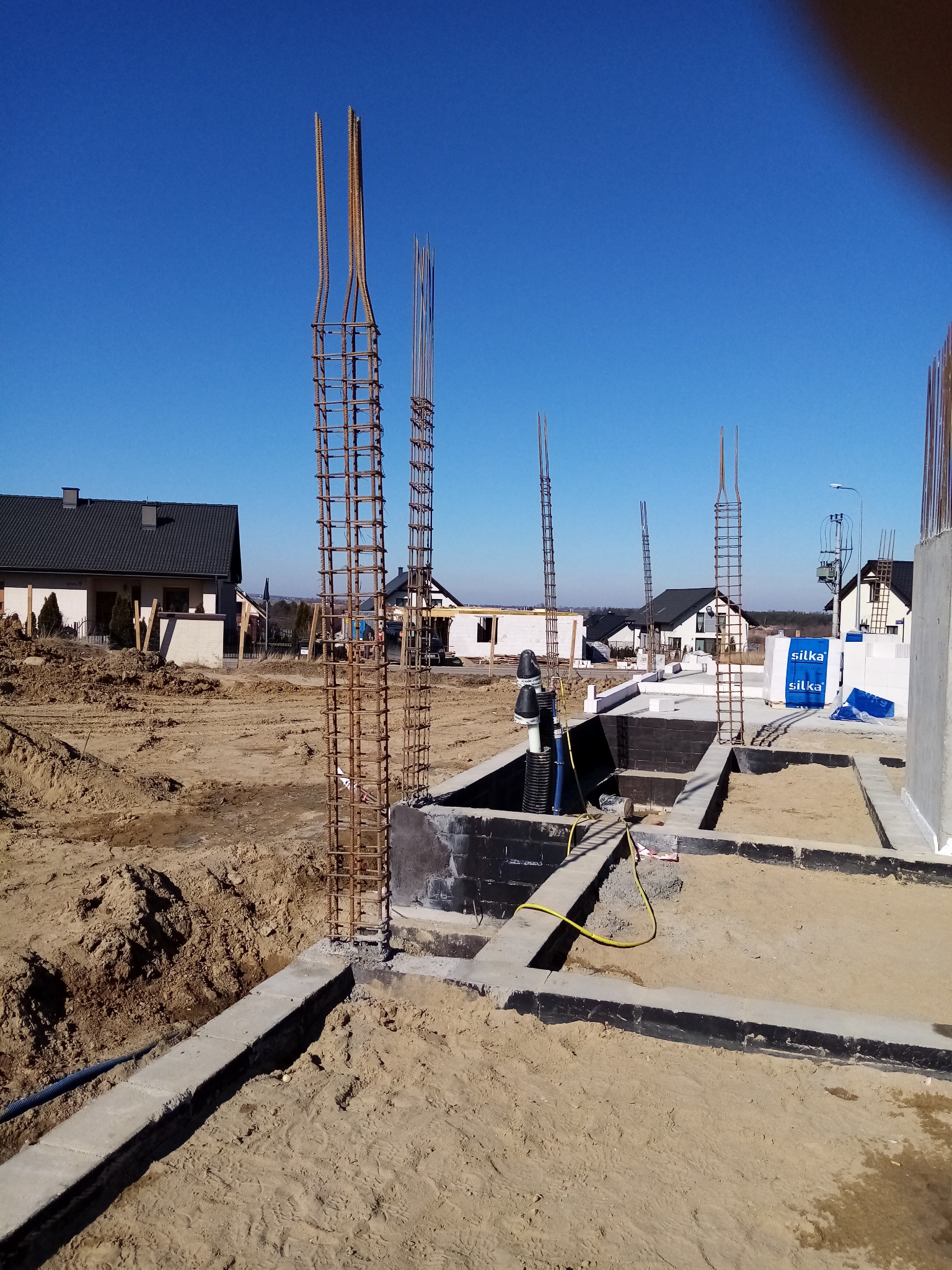 Fundamenty domu jednorodzinnego w trakcie budowy, widoczne zbrojenia i izolacja przeciwwodna, z boku składowane bloczki Silka.