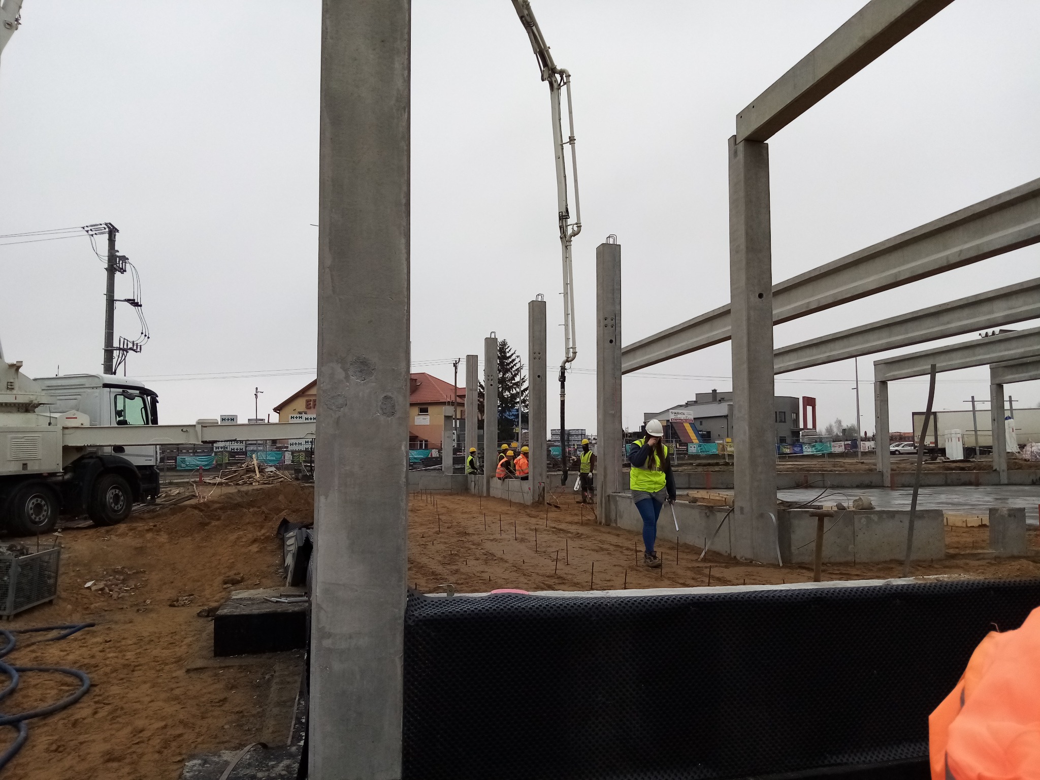 Montaż prefabrykowanych słupów żelbetowych na placu budowy, widoczna pompa do betonu i pracownicy w odblaskowych kamizelkach.