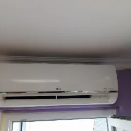 Biały klimatyzator LG Dual Inverter zamontowany na fioletowej ścianie nad oknem, widoczny wyświetlacz temperatury 20 stopni Celsjusza.