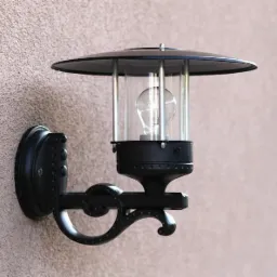 Czarny, metalowy kinkiet zewnętrzny z przezroczystym kloszem i żarówką, zamontowany na teksturowanej ścianie budynku.