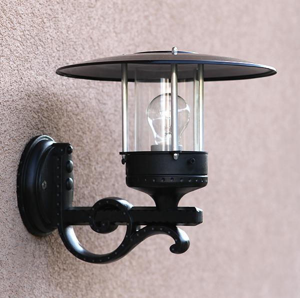 Czarny, metalowy kinkiet zewnętrzny z przezroczystym kloszem i żarówką, zamontowany na teksturowanej ścianie budynku.