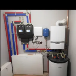 Zmontowana instalacja grzewcza z piecem gazowym Vaillant, zasobnikiem wody i rurami w kolorach czerwonym i niebieskim, widoczna na tle szarej ściany.