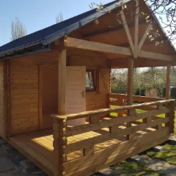 Drewniany domek letniskowy z otwartymi drzwiami, częściowo zadaszonym tarasem i czarnym dachem, posadowiony na betonowych bloczkach w ogrodzie.