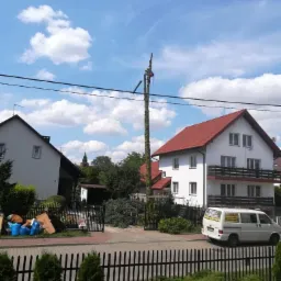 Pracownik w kasku i uprzęży, na wysokim pniu drzewa w trakcie wycinki, z widocznymi fragmentami ściętych gałęzi i budynkami w tle.