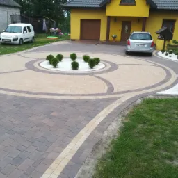 Ułożona z kostki brukowej dwukolorowa nawierzchnia podjazdu do żółtego domu jednorodzinnego, z okrągłym wzorem i rabatą z białych kamieni i kulistych krzewów pośrodku. Na podjeździe zaparkowany...