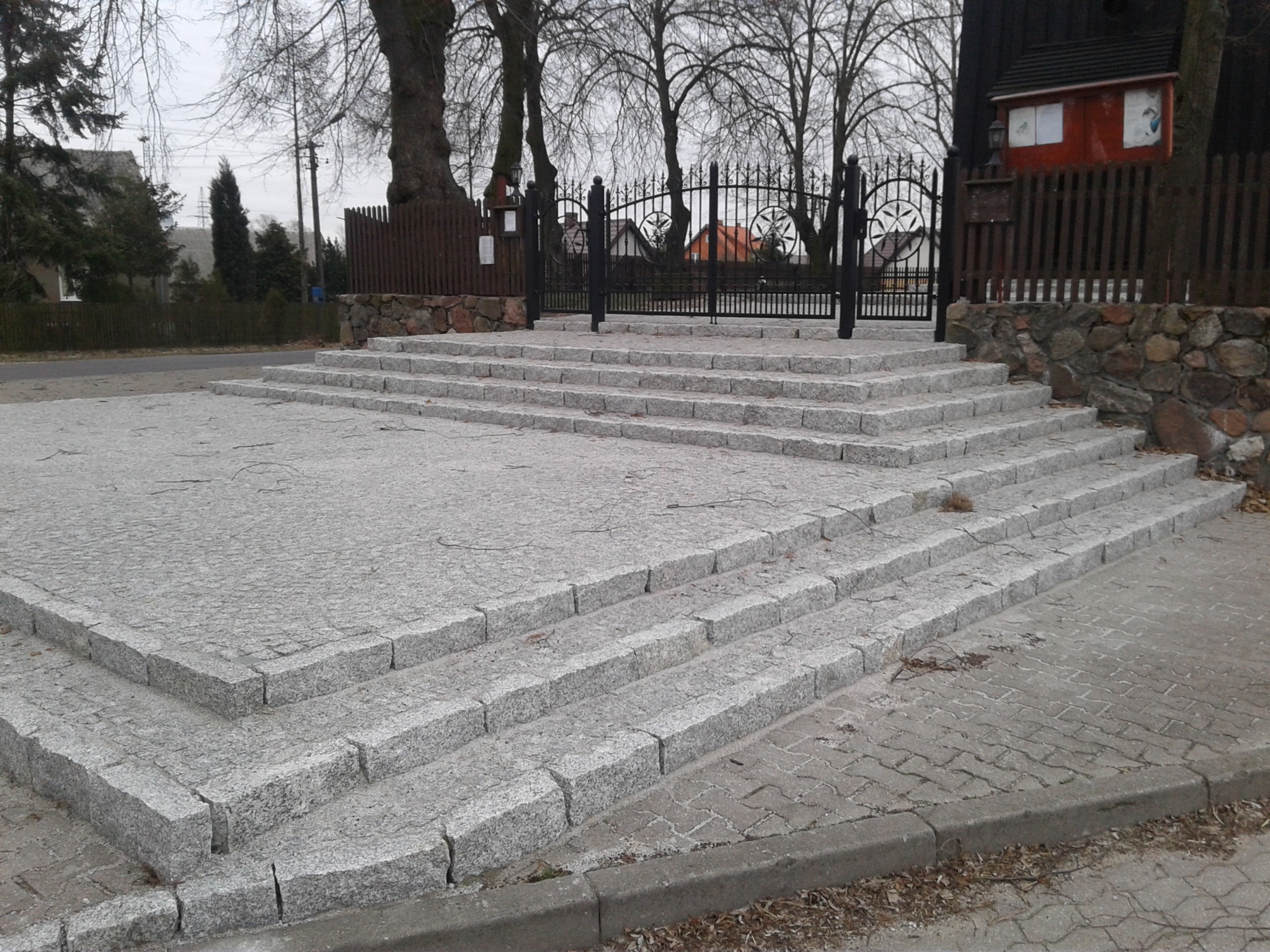 Szerokie, granitowe schody prowadzące do ozdobnej, metalowej bramy, otoczone kamiennym murem i drewnianym ogrodzeniem, z widocznymi drzewami bez liści w tle.