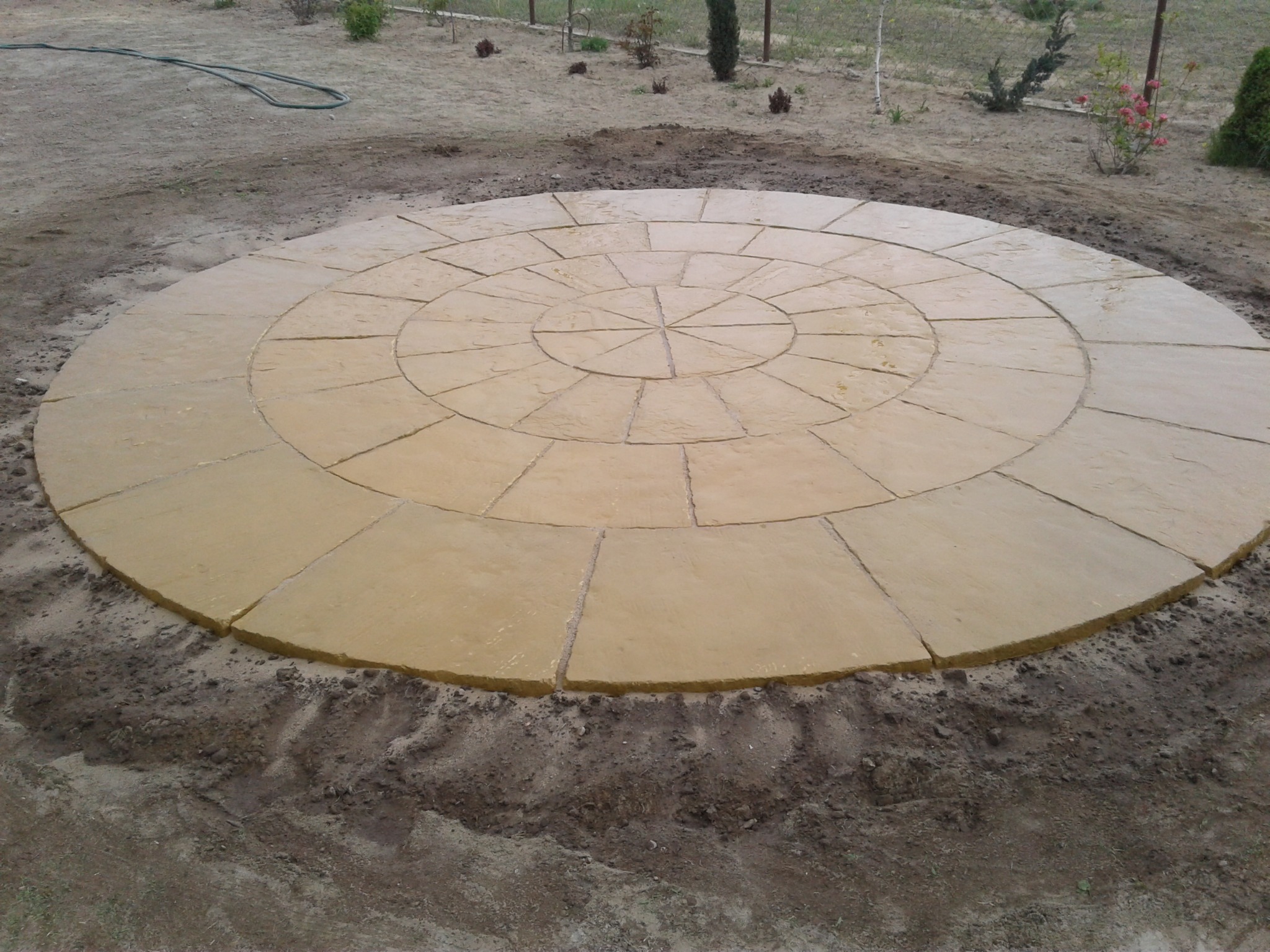 Okrągły plac z żółtych płyt kamiennych ułożonych w koncentryczny wzór, otoczony ziemią, w tle trawnik z młodymi drzewkami.