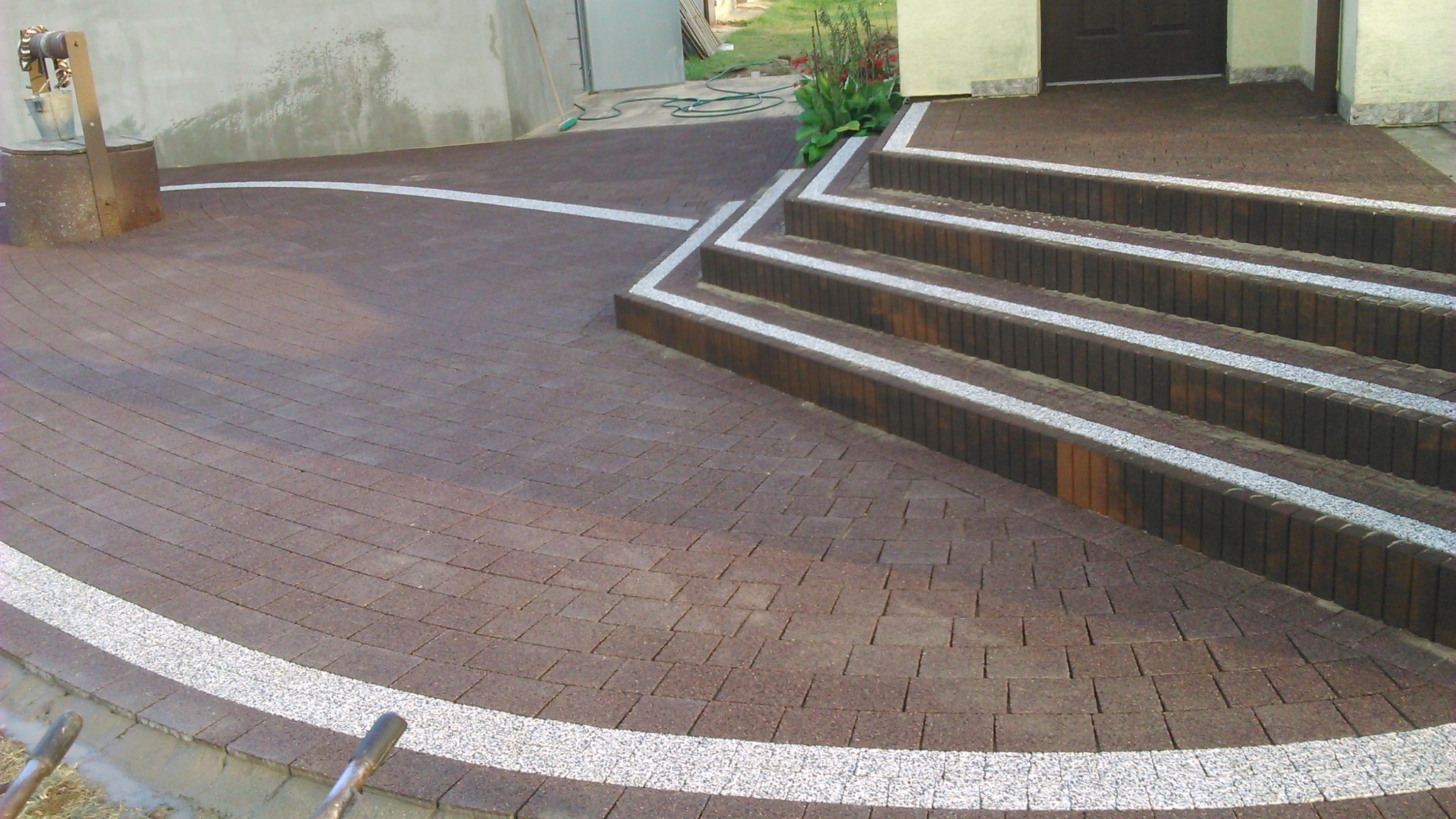 Brązowa kostka brukowa ułożona w łukowaty wzór z białymi akcentami, prowadząca do schodów wejściowych z kontrastującymi białymi wypełnieniami. Stara studnia z wiadrem po lewej stronie.