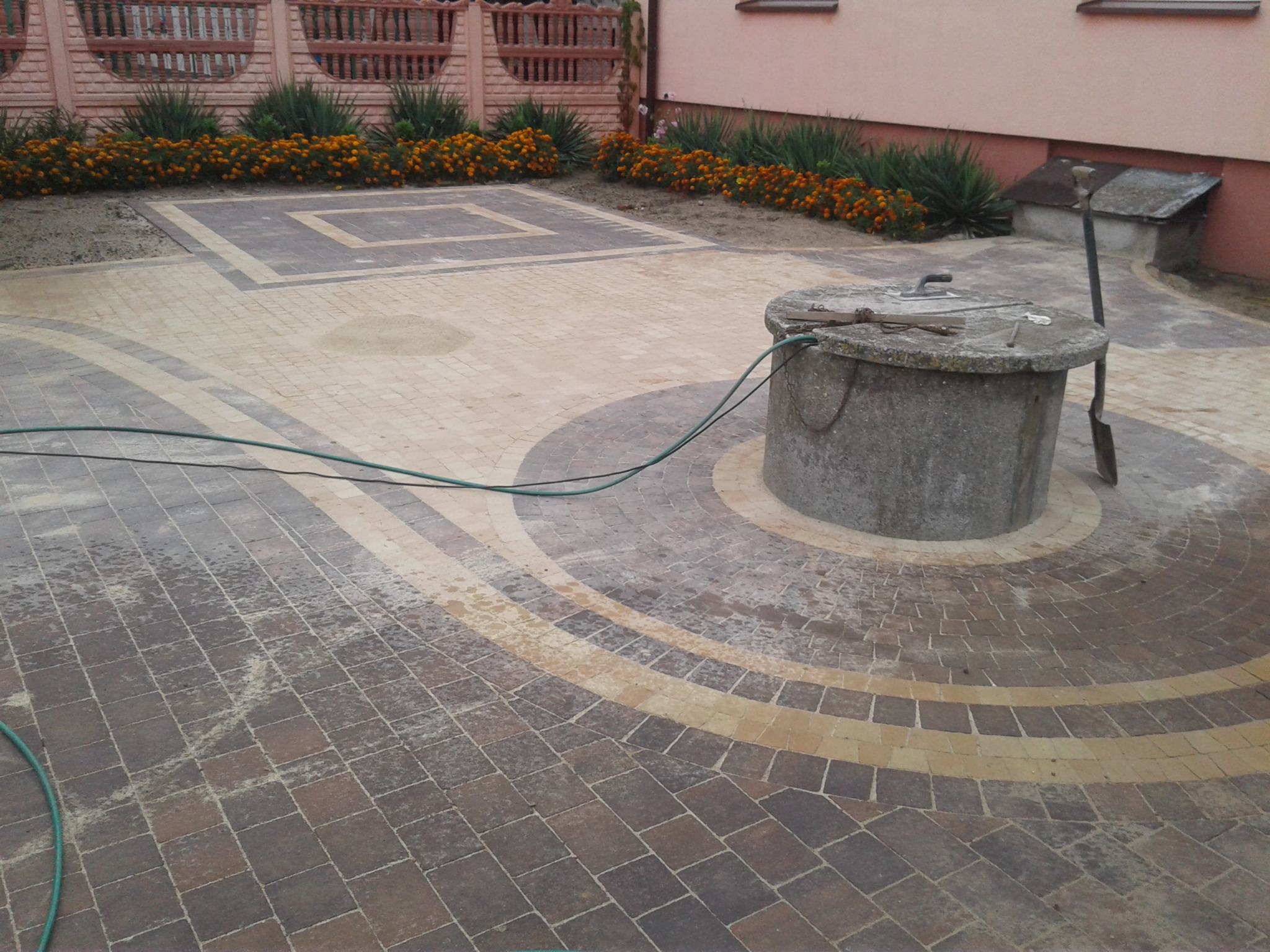 Ułożona kostka brukowa w dwóch odcieniach wokół betonowej studni, z widocznymi śladami wody i narzędziami na studni, otoczona rabatami z pomarańczowymi kwiatami i ozdobnym ogrodzeniem.