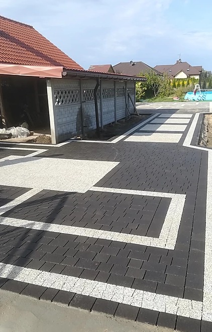 Ukończony podjazd z ciemnej kostki brukowej z jasnymi wstawkami, tworzący geometryczny wzór przed garażem z czerwonym dachem i betonowym ogrodzeniem.