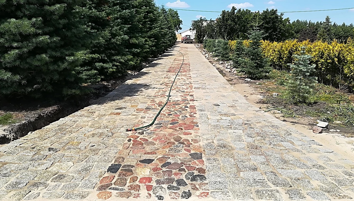 Długa, wybrukowana droga z kostki granitowej z pasem czerwono-czarnych kamieni pośrodku, otoczona zielonymi drzewami i krzewami, z traktorem w oddali i wężem ogrodowym na ścieżce.