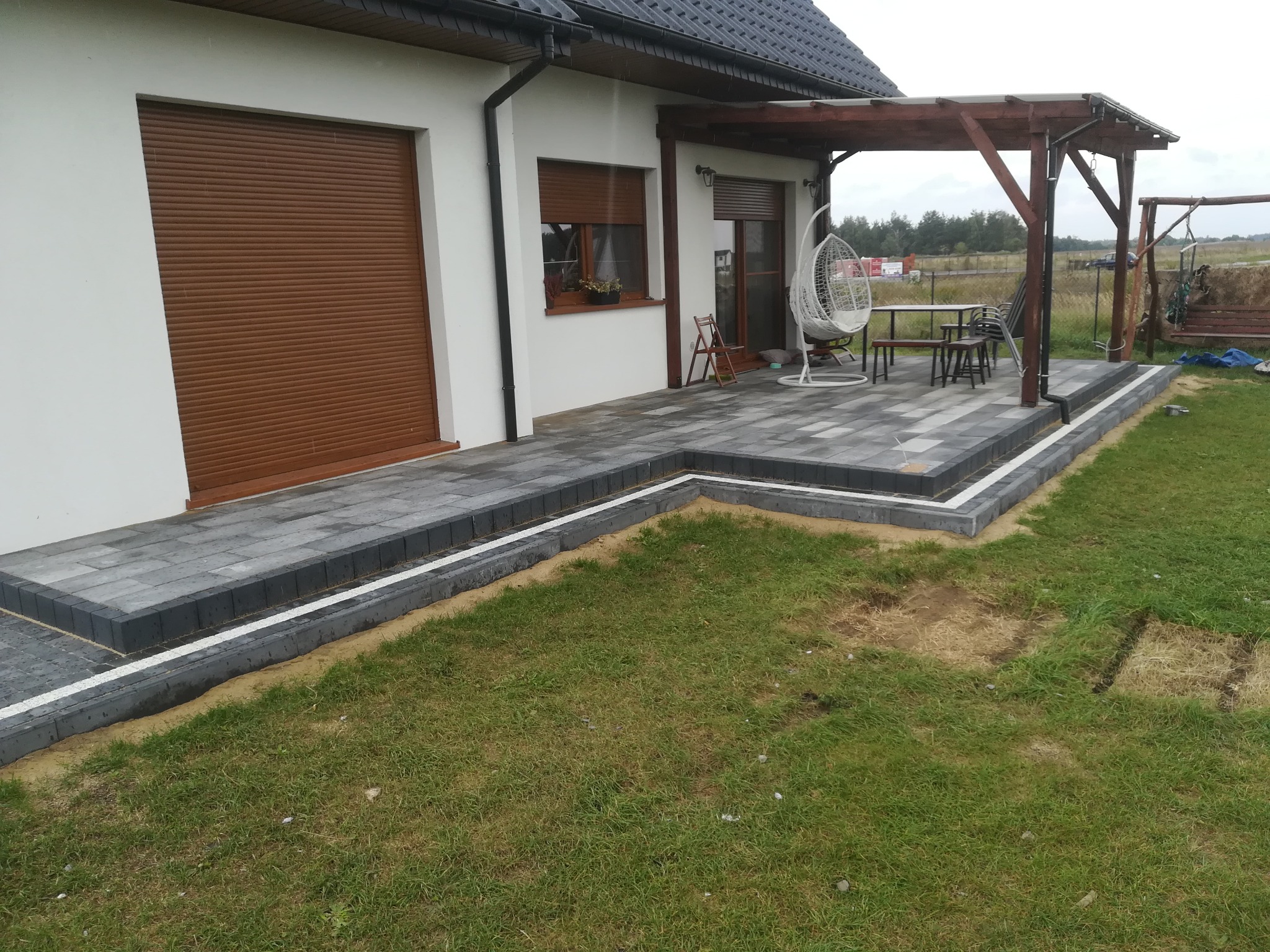Szara kostka brukowa ułożona na tarasie przylegającym do białego domu z brązowymi roletami, widoczna pergola z drewnianą konstrukcją i wiszącym fotelem, trawnik z uszkodzeniami.