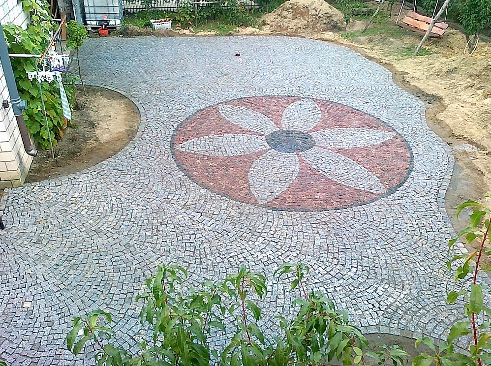Dziedziniec wyłożony szarą kostką z dekoracyjnym wzorem kwiatu w odcieniach czerwieni i szarości, widoczne przygotowane podłoże i roślinność.