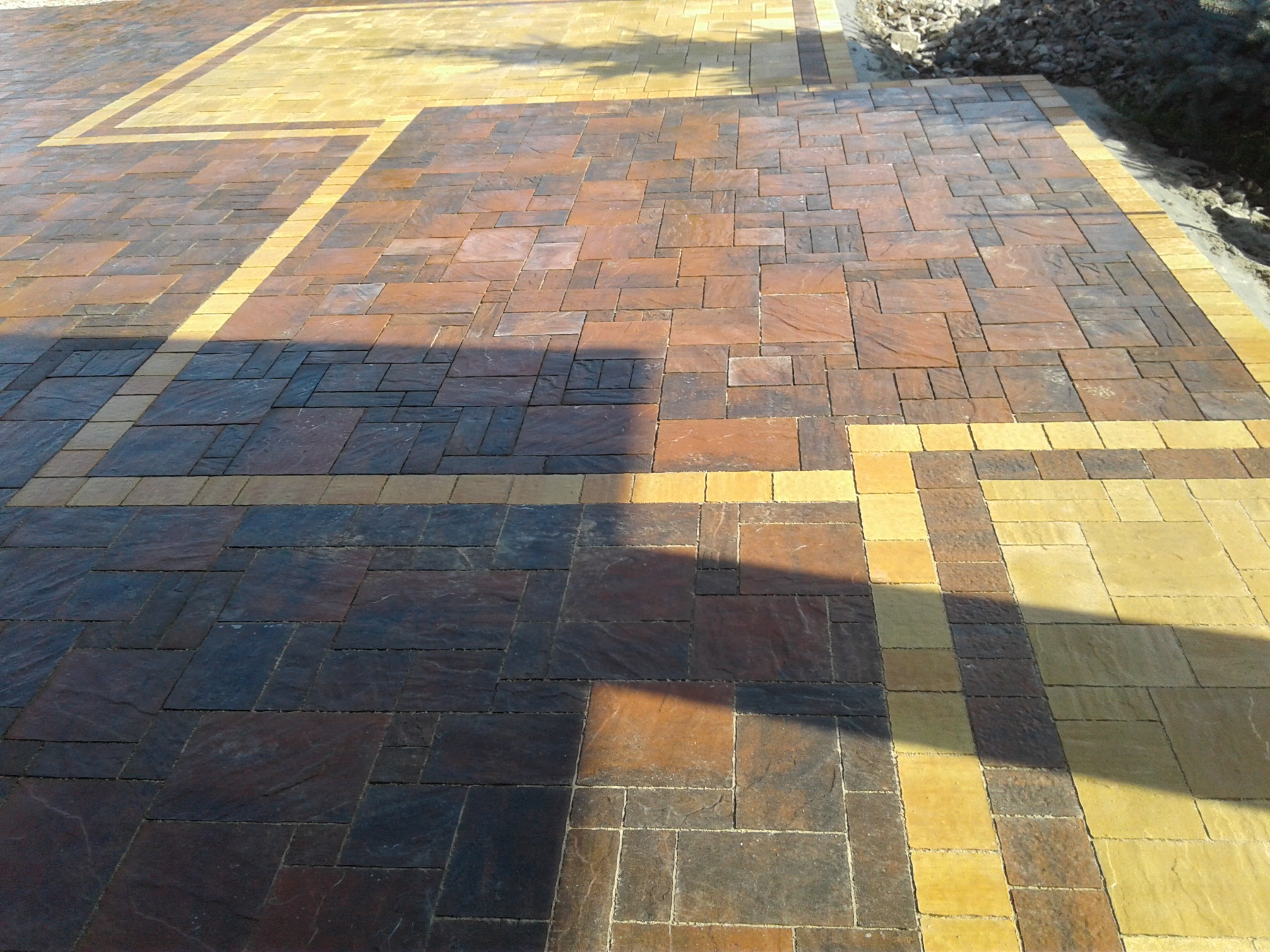 Ułożona nawierzchnia z kostki brukowej w odcieniach brązu i żółci, z widocznym cieniem padającym na fragment powierzchni. Różne rozmiary kostek tworzą geometryczny wzór.