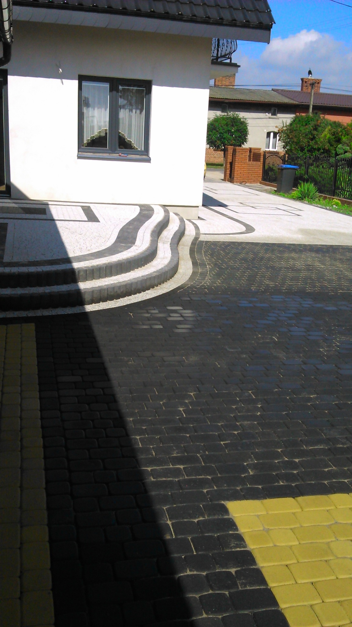 Nowoczesny podjazd z ciemnej kostki brukowej z żółtymi akcentami, prowadzący do domu z białymi ścianami i schodami o falistym kształcie z jasnej kostki.