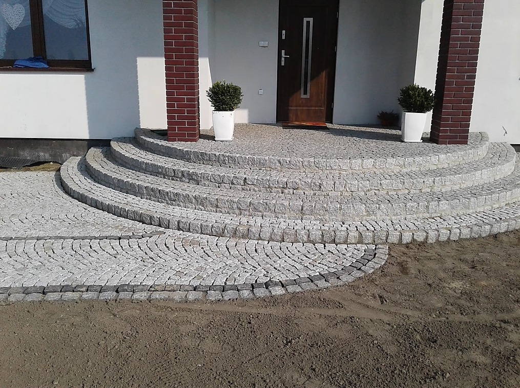 Półokrągłe schody wejściowe z jasnoszarej kostki granitowej prowadzące do domu z ceglanymi filarami i białymi donicami z roślinami.