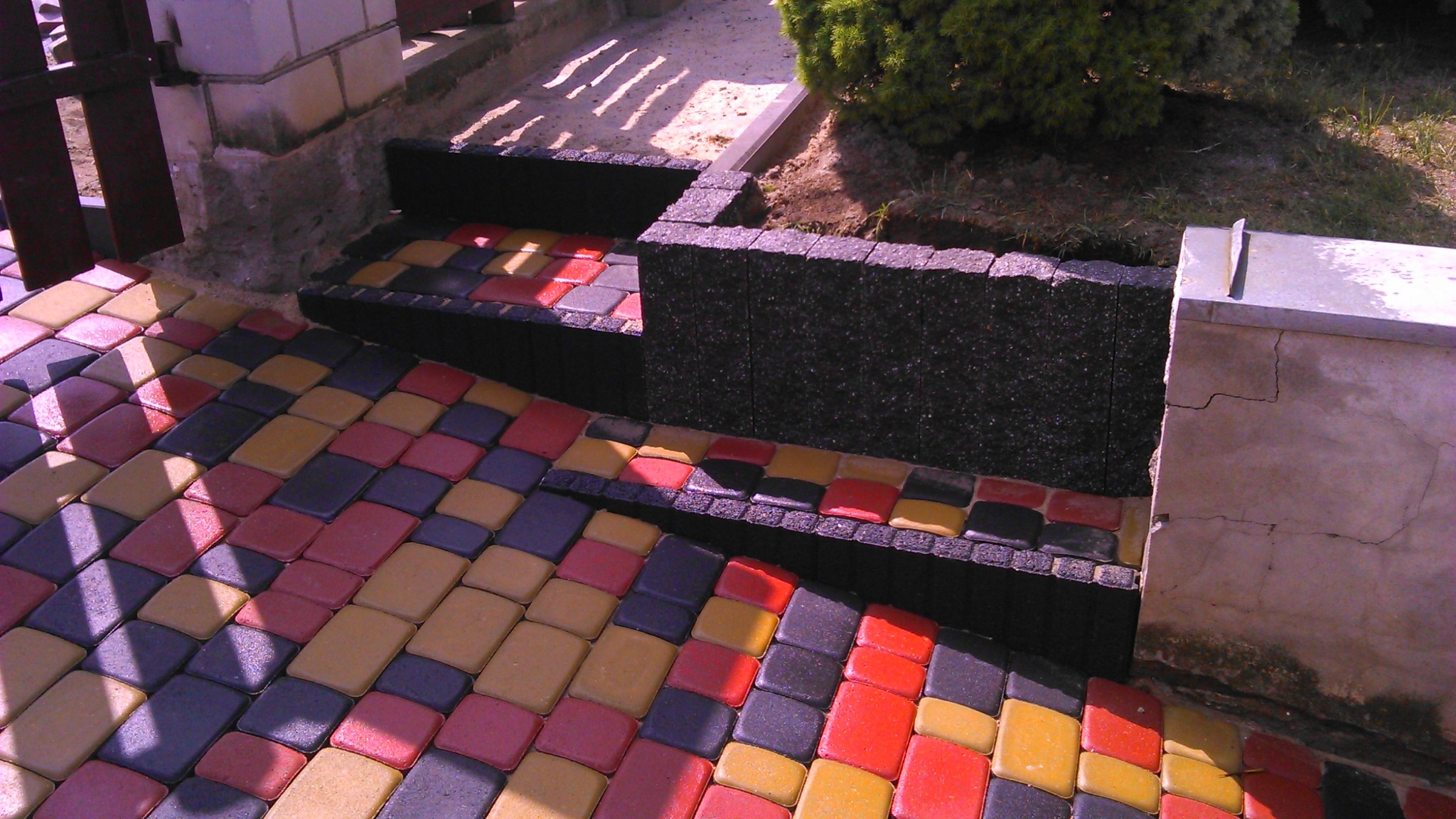 Kolorowa kostka brukowa ułożona na podjeździe, tworząca schody z czarnymi obrzeżami, z widocznym fragmentem trawnika i krzewu.