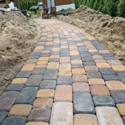 Ścieżka z wielobarwnej kostki brukowej prowadząca przez ogród, ubijarka wibracyjna w tle, świeżo usypana ziemia po bokach.